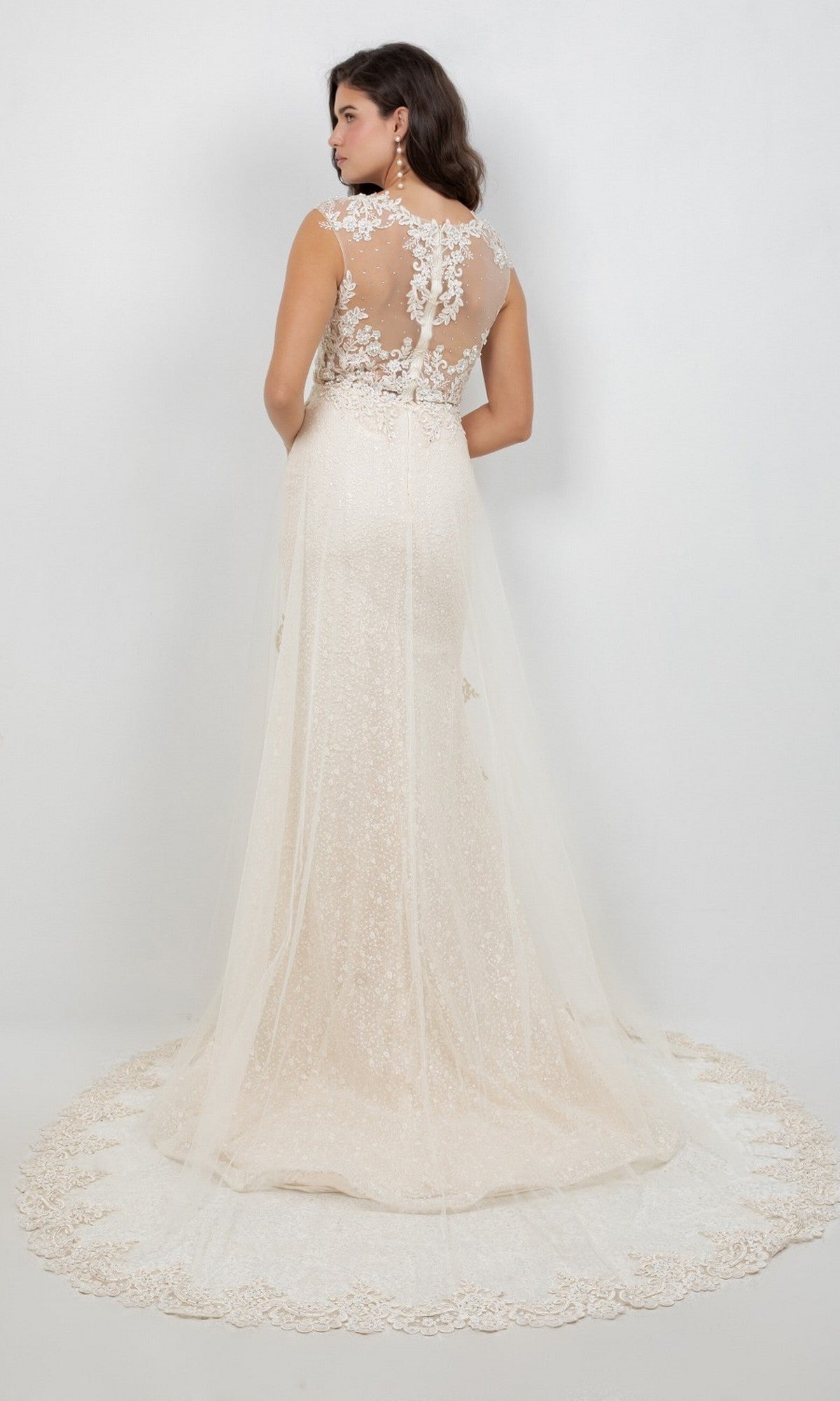 A woman stands facing away, wearing the Long Formal Dress: Cinderella Couture 8506J—a white, floor-length gown crafted from delicate tulle with lace detailing and a sheer back—set against a plain white background.